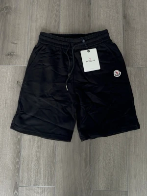 Svarta shorts från Moncler med snörning - Snygga svarta shorts från Moncler med elastisk midja och snörning. Klassisk Moncler-logga på vänster ben och mjukt material som känns skönt mot huden. Perfekta för en avslappnad och sportig stil.