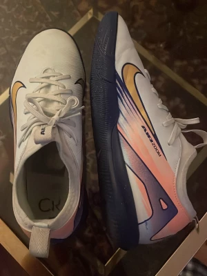Nike Air Zoom vita fotbollsskor  - Nike Air Zoom fotbollsskor i vitt med blå och orange detaljer samt guld Swoosh på sidan. Skorna har snörning, blå yttersula och en modern design med inslag av rosa och marinblått. Materialet är en mix av syntet och textil, perfekt för dig som vill sticka ut på planen.