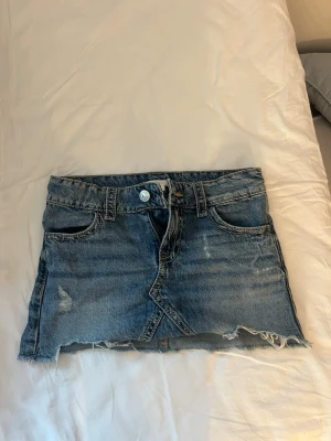 Blå jeanskjol  - Kort jeanskjol i blå denim med slitna detaljer och rå, fransig kant nedtill. Kjolen har klassiska fem fickor, bälteshällor och knappgylf. Perfekt för en avslappnad och trendig look.
