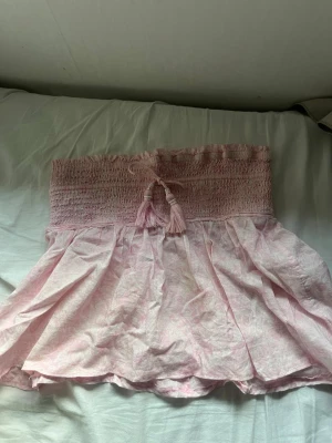 Ljusrosa shorts med smock och tofsar - Säljer ett par ljusrosa shorts med bred smock i midjan och dekorativa tofsar framtill. Shortsen har en luftig och flowy passform, perfekta för varma dagar. Materialet känns lätt och bekvämt mot huden. Kjolen var lång men jag har sytt om den till kort, eftersom att jag tyckte den blev för liten. Den har några fläckar som man knappt ser. Kan budda väldigt låg eftersom att jag bara vill bli av med sakerna!