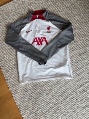 Extremt snygg Nike x Liverpool träningströja vit/grå - Snygg långärmad träningströja från Nike med Liverpool FC-logga och AXA sponsortryck. Tröjan är vit med grå ärmar och röd krage, har halv dragkedja och är tillverkad i Dri-FIT material som andas. Perfekt för fotbollsträning eller gym.