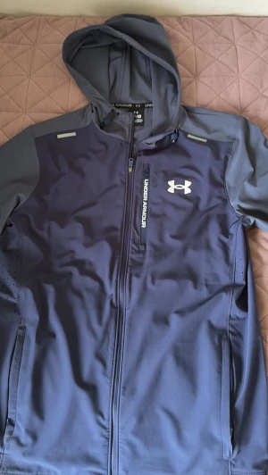 Blå hoodie från Under Armour - Snygg blå hoodie från Under Armour med dragkedja framtill och huva. Har en bröstficka med dragkedja och vit logga på bröstet. Tillverkad i ett sportigt, stretchigt material som passar perfekt till träning eller chill. Under Armour-tryck längs nederkanten bak.