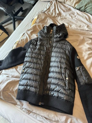 Svart pufferjacka från Moncler - Snygg svart pufferjacka från Moncler med stickade ärmar och detaljer. Jackan har huva, dragkedja framtill, två fickor med tryckknappar och en extra ficka med dragkedja på ärmen. Perfekt för kyliga dagar och riktigt trendig look.