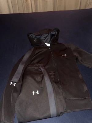 Svart set från Under Armour - Snyggt svart loungewear-set från Under Armour med matchande hoodie och byxor. Setet har dragkedja, huva med snörning och diskreta loggor på bröstet och benet. Materialet är mjukt och stretchigt, perfekt  passar xs 