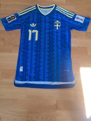 Blå Sverige fotbollströja Adidas s - Säljer en blå fotbollströja från Adidas med Sveriges landslagsemblem, gula detaljer och nummer 17 på både fram- och baksida. Tröjan har trycket 'GYÖKERES' på ryggen, korta ärmar och ett unikt mönster i tyget. Perfekt för match eller träning. Strl s och det är playerversion. Detta är en replika. Prefekt för att suport sverige i fotbolls vm