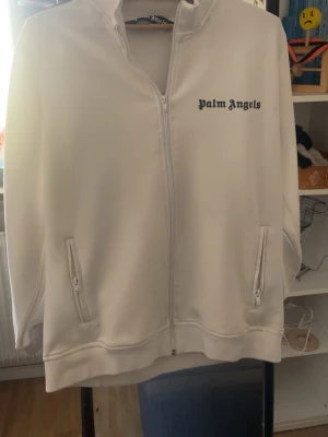 Vit ziptröja från Palm Angels - Snygg vit ziptröja från Palm Angels med svart logga på bröstet. Tröjan har dragkedja framtill, två fickor med dragkedja och svarta ränder längs ärmarna. Tillverkad i ett mjukt och stretchigt material som ger en sportig vibe.