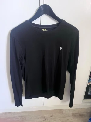 Svart långärmad T-shirt Polo Ralph Lauren - Svart långärmad T-shirt  från Polo Ralph Lauren med rund halsringning och den klassiska broderade loggan på bröstet. Enkel och stilren design som passar till det mesta. Tillverkad i mjuk bomull för en skön känsla.