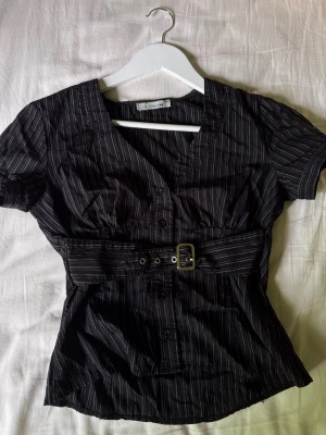 Svart blus med bälte och ränder - Snygg svart blus från Bik Bok med tunna vita ränder och markerad midja med bälte. Blusen har korta ärmar, knappar framtill och en feminin v-ringning. Perfekt för en trendig och stilren look.