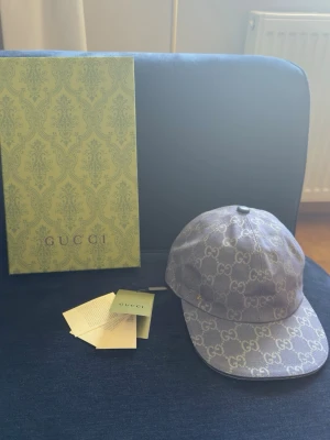 Gucci keps i blått monogram - Säljer en snygg keps från Gucci i grått tyg med det klassiska GG-monogrammet över hela kepsen. Kepsen har böjd skärm och justerbar rem bak. Perfekt för dig som vill ha en exklusiv och trendig accessoar till din outfit.