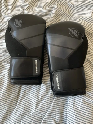 Svarta boxningshandskar Hayabusa - Säljer ett par svarta boxningshandskar från Hayabusa med snygga grå detaljer och logga på ovansidan. Handskarna har breda kardborreband för extra stöd och är tillverkade i slitstarkt syntetmaterial. Perfekta för träning och sparring.