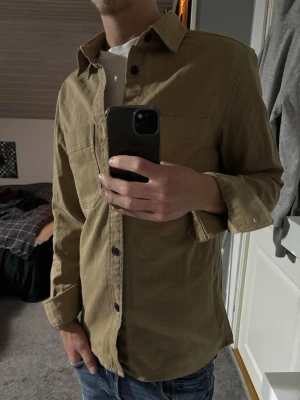 Overshirt  - Snygg beige overshirt med klassisk krage och svarta knappar framtill. Skjortan har lång ärm och bröstficka, perfekt att bära öppen över en t-shirt. Stilren och enkel design som passar till jeans.