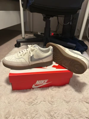 Nike skor  - Säljer ett par fräscha vita Nike sneakers med grå swoosh och beigegrå sula. Skorna har klassisk låg profil, perforerad tåbox och snörning. Passar perfekt till streetwear och har en tidlös look som funkar till det mesta.