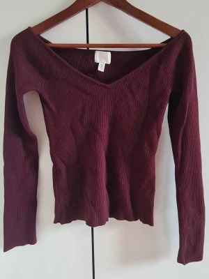 Vinröd ribbad långärmad topp - Snygg vinröd ribbad topp med v-ringning och långa ärmar. Toppen har en figurnära passform och är tillverkad i ett mjukt stretchigt material som sitter skönt på kroppen. Perfekt att styla med jeans eller kjol för en trendig look.