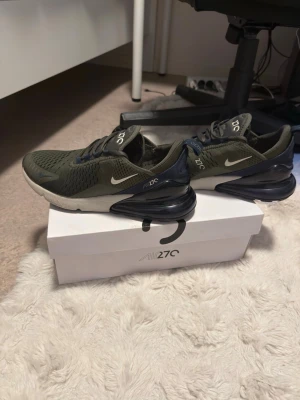 Nike Air Max 270  - Nike Air Max 270 sneakers i olivgrönt mesh med svarta och vita detaljer. Skorna har en stor synlig Air-enhet i hälen, vita swoosh-loggor och texten 'AIR 270' på plösen. Lågt skaft och snörning med mörkblå öglor. Perfekta för dig som gillar streetwear.