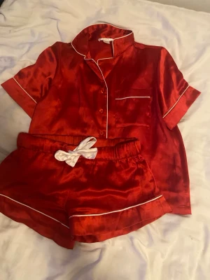 Röd satinpyjamas med vita detaljer - Snygg röd pyjamas i glansigt satinmaterial med kortärmad skjorta och matchande shorts. Setet har vita kontrastkanter och en vit rosett i midjan på shortsen. Perfekt för mysiga kvällar hemma.