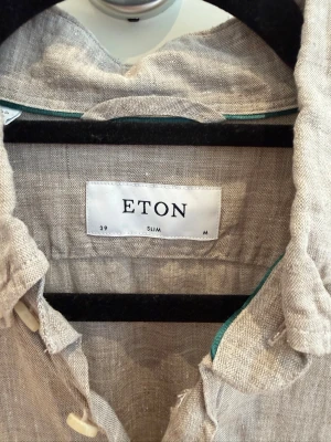 Eton Linneskjorta – Slim Fit, Strl 39 – Nypris ca 2 000 kr - Snygg och stilren linneskjorta från Eton i en elegant brun/ljusbrun nyans. Tillverkad i 100 % linne av hög kvalitet, vilket ger en luftig och bekväm känsla – perfekt för både sommarens varma dagar och mer uppklädda tillfällen.  ✔ Storlek 39 Slim Fit ✔ 100 % linne ✔ Button down-krage ✔ Praktisk bröstficka ✔ Tidlös och elegant design ✔ Kanonfint skick  Nypris cirka 2 000 kr.  Skjortan är väl omhändertagen och kommer från ett djur- och rökfritt hem.  Ett perfekt val för dig som uppskattar kvalitet.