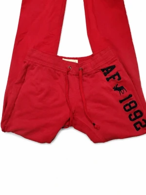 Röda mjukisbyxor Abercrombie & Fitch - Snygga röda mjukisbyxor från Abercrombie & Fitch med svart broderad text 'AF 1892' och älg på ena benet. Byxorna har dragsko i midjan och sidofickor. Perfekta för chill dagar och riktigt sköna tack vare mjukt bomullsmaterial.