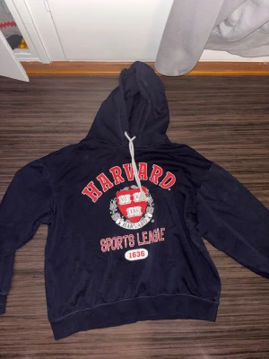 Svart Harvard hoodie med tryck - Snygg svart hoodie med stor huva och vitt snöre ifrån H&M. Framtill finns ett stort Harvard-tryck i rött och vitt samt texten 'Sports League 1636'. Hoodien har ribbade muddar och är gjord i mjukt bomullsmaterial. 