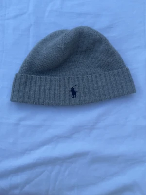 Ralph lauren mössa - Snygg grå ribbstickad mössa från Polo Ralph Lauren med klassisk broderad logga framtill. Tillverkad i mjukt material som håller dig varm och stilren under kalla dagar. Perfekt accessoar för en clean och avslappnad look. Hör av er vid frågor och funderingar. Mössan har inga defekter