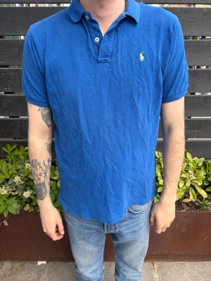 Blå pikétröja från Ralph Lauren - Klassisk blå pikétröja från Ralph Lauren med korta ärmar och den ikoniska broderade loggan på bröstet. Tröjan har krage och knappar vid halsen. Tillverkad i mjuk bomull som känns skön mot huden. Perfekt till jeans eller shorts.