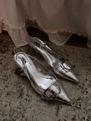Silvriga pumps med spänne från Zara - Säljer ett par silvriga kitten heels från Zara med spetsig tå och låg klack. Skorna har en stor dekorativ metallspänne framtill och smal rem runt hälen. Tillverkade i ett glansigt material.