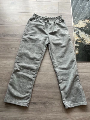 Grå mjukisbyxor med broderad logga - Säljer ett par grå mjukisbyxor med resår i midjan och två sidofickor. Byxorna har en vit broderad logga på vänster sida och är tillverkade i mjukt sweatshirtmaterial. Perfekta för chill dagar eller när du vill vara bekväm. Den är stor för att vara L