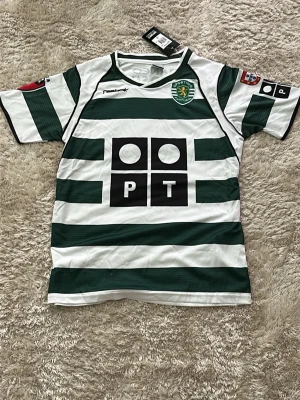 Sporting CP matchtröja Reebok grön/vit - Säljer en officiell Sporting CP matchtröja från Reebok i grönt och vitt med breda horisontella ränder. Tröjan har klubbmärke på bröstet, sponsortryck och korta ärmar. Perfekt för fotbollsfans som vill visa sitt stöd för laget.