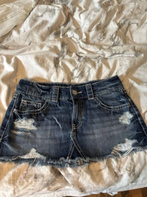 Blå jeanskjol med slitningar - Snygg kort jeanskjol i blå denim med slitna detaljer och fransig nederkant. Klassisk femficksmodell med vita kontrastsömmar och knappstängning framtill. Perfekt för en avslappnad och trendig look. Bara använd en gång och passar xs och S! 