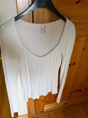 Vit långärmad topp från H&M - Säljer en enkel och stilren vit långärmad topp. Toppen har en djup v-ringning med en söt spetskant runt halsen. Känns skön mot huden. Perfekt att matcha med jeans eller kjol.