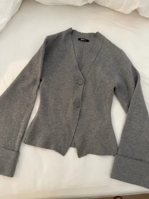 Grå kofta med vida ärmar från Gina  - Snygg grå kofta från Gina tricot med två stora knappar framtill och vida ärmar med breda uppvik och dekorativa knappar. Koftan har en figurnära passform och är tillverkad i mjukt stickat material. Perfekt för lager-på-lager-stil. Storlek xs, som helt ny.