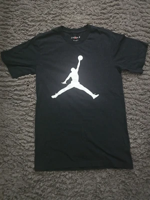 Svart Jordan t-shirt med logga - Svart t-shirt från Jordan med den klassiska vita Jumpman-loggan på bröstet. T-shirten har rund halsringning och korta ärmar. Perfekt för dig som gillar sportig och stilren streetwear.