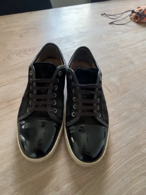 Lanvins - Snygga svarta lanvins med glansiga lackdetaljer på tån och klassisk vit sula. Skorna har svarta snören och metallöljetter för en stilren look. Perfekta för dig som vill ha en enkel men ändå trendig sneaker till vardags. Finns även box
