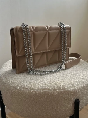 Beige quiltad axelväska med kedja - Stilren beige axelväska i quiltat mönster med silverfärgad kedjerem och magnetlås. Väskan har en rektangulär form och är tillverkad i ett slätt, läderliknande material. Perfekt för att lyfta din outfit med en trendig touch.