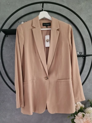 Beige kavaj från KappAhl - Stilren beige kavaj från KappAhl med klassisk krage och enkel knappstängning. Kavajen har raka fickor framtill och är tillverkad i ett mjukt material som ger en snygg passform. Perfekt för dig som vill ha en clean och modern look.