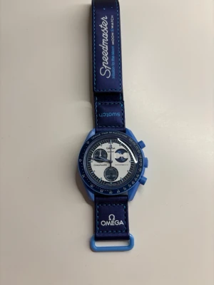 Omega x Swatch blå MoonSwatch - Snygg Omega x Swatch MoonSwatch med blå boett och blått band. Klockan har vit urtavla med blå detaljer och flera subdials. Bandet är dekorerat med Speedmaster- och Omega-loggor. Perfekt för dig som gillar ikoniska samarbeten och cool design.