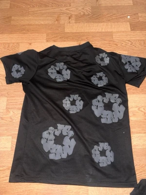 Svart t-shirt från Indenimtears - Cool svart t-shirt från Indenimtears med unika grå blommönster tryckta över hela tröjan. T-shirten har korta ärmar och en klassisk rund halsringning. Perfekt för dig som vill sticka ut med en originell och trendig stil.