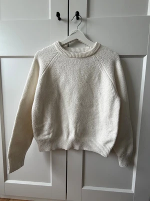 Klassisk vit stickad tröja - En enkel och stilren vit stickad tröja med rund hals och raglanärmar. Tröjan har ribbstickade muddar vid ärmslut, hals och nederkant. Perfekt för lager-på-lager och passar till många olika outfits. Materialet ser ut att vara mjukt och värmande.