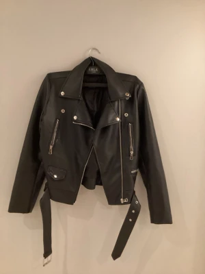 Svart biker skinnjacka med bälte konstläder - Snygg svart skinnjacka i klassisk bikerstil från Serla. Jackan har silvriga dragkedjor, snedställd frontzip, fickor och ett bälte i midjan. Perfekt för dig som vill ha en edgy och tidlös look. Passar till allt och ger en cool vibe.