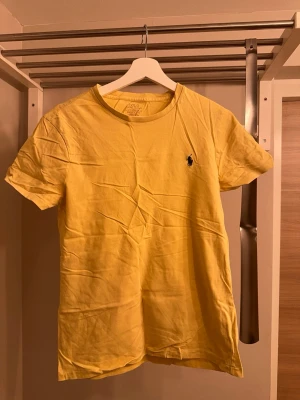 Gul t-shirt från Polo Ralph Lauren - Snygg gul t-shirt från Polo Ralph Lauren med klassisk rund hals och den ikoniska loggan broderad på bröstet. Tillverkad i mjuk bomull och har en normal passform. Perfekt för en avslappnad och stilren look.