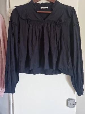 Svart volangblus med lång ärm - Svart blus med volangdetaljer och v-ringning. Långärmad modell med rynkade partier och spetsinslag vid ärmarna. Lös passform och lätt, vävt material. Perfekt för en trendig och avslappnad look.