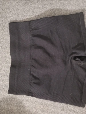 Svarta shaping shorts med hög midja - Säljer ett par svarta shaping shorts med hög midja och ribbade kanter. De är sömlösa och har extra rynkning bak för att framhäva formen. Materialet är stretchigt och bekvämt, perfekt för att skapa en slät siluett under kläder.
