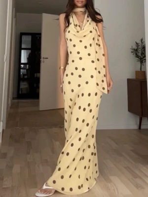 Lång prickig klänning med scarfdetalj - Säljer en lång, ljusgul klänning med bruna prickar och scarfdetalj vid halsen. Klänningen har öppen rygg och en lös, elegant passform. Perfekt för dig som vill sticka ut med ett unikt mönster och snygg siluett.