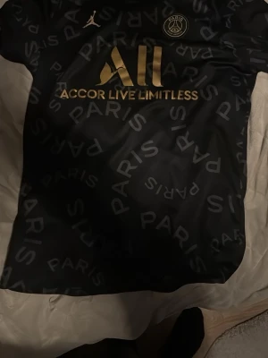 Svart PSG x Jordan t-shirt - Snygg svart t-shirt från PSG och Jordan med guldiga detaljer och texten 'Accor Live Limitless' på bröstet. T-shirten har ett mönster med ordet 'PARIS' över hela tyget och PSG-loggan på bröstet. Perfekt för dig som gillar fotboll och streetwear.