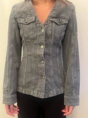 Grå figursydd jeansjacka med nitar - Helt ny snygg grå jeansjacka med figurnära passform och dekorativa nitar längs ärmarna. Jackan har bröstfickor med lock och knappar framtill. Storlek XS men passar S med❣️ Sista bilden är inte min)