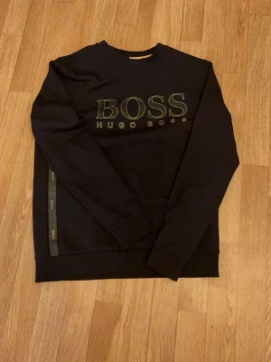Svart sweatshirt från Hugo Boss - Snygg svart sweatshirt från Hugo Boss med stort logotryck i gult på bröstet och dekorativt band med BOSS-tryck längs sidan. Klassisk rund halsringning och ribbade muddar vid ärmslut och nederkant. Perfekt för en stilren och avslappnad look.