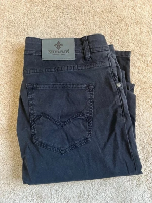 Mörkblå slim fit jeans från Morris - Snygga mörkblå jeans från Morris med slim fit passform och klassisk femficksdesign. Jeansen har en diskret logga på bakfickan och patch med Morris-märke i midjan. Tillverkade i ett mjukt och stretchigt bomullsmaterial för extra komfort.