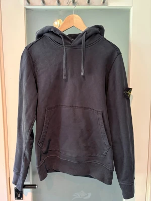 Mörkgrå hoodie från Stone Island - Säljer en mörkgrå hoodie från Stone Island med klassisk patch på vänster ärm. Hoodien har dragsko i huvan, känguruficka framtill och ribbade muddar vid ärmslut och nederkant. Perfekt för en avslappnad streetwear-look.