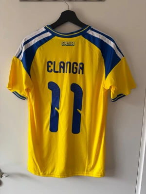 Sverige matchtröja Elanga #11 - Gul och blå Sverige matchtröja från Adidas med Elanga och nummer 11 på ryggen. Tröjan har korta ärmar, blå detaljer på axlar och ärmslut samt broderad landslagssköld och Adidas-logga på bröstet.