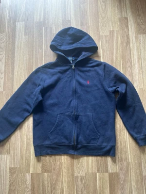 Mörkblå hoodie från Polo Ralph Lauren - Mörkblå hoodie med dragkedja från Polo Ralph Lauren. Klassisk design med huva, känguruficka och det ikoniska röda Polo-logot på bröstet. Tillverkad i mjukt bomullsmaterial som är skönt att ha på sig. Perfekt för en avslappnad och stilren look.