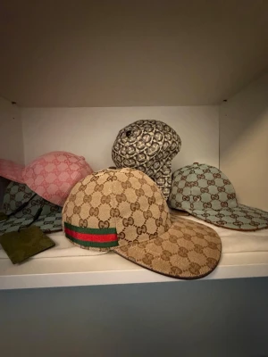 Beige Gucci keps med GG-mönster - Säljer en beige keps från Gucci med det klassiska GG-monogrammet över hela kepsen. På sidan finns en grön och röd rand som ger en ikonisk touch. Kepsen har böjd skärm och är tillverkad i canvasmaterial.